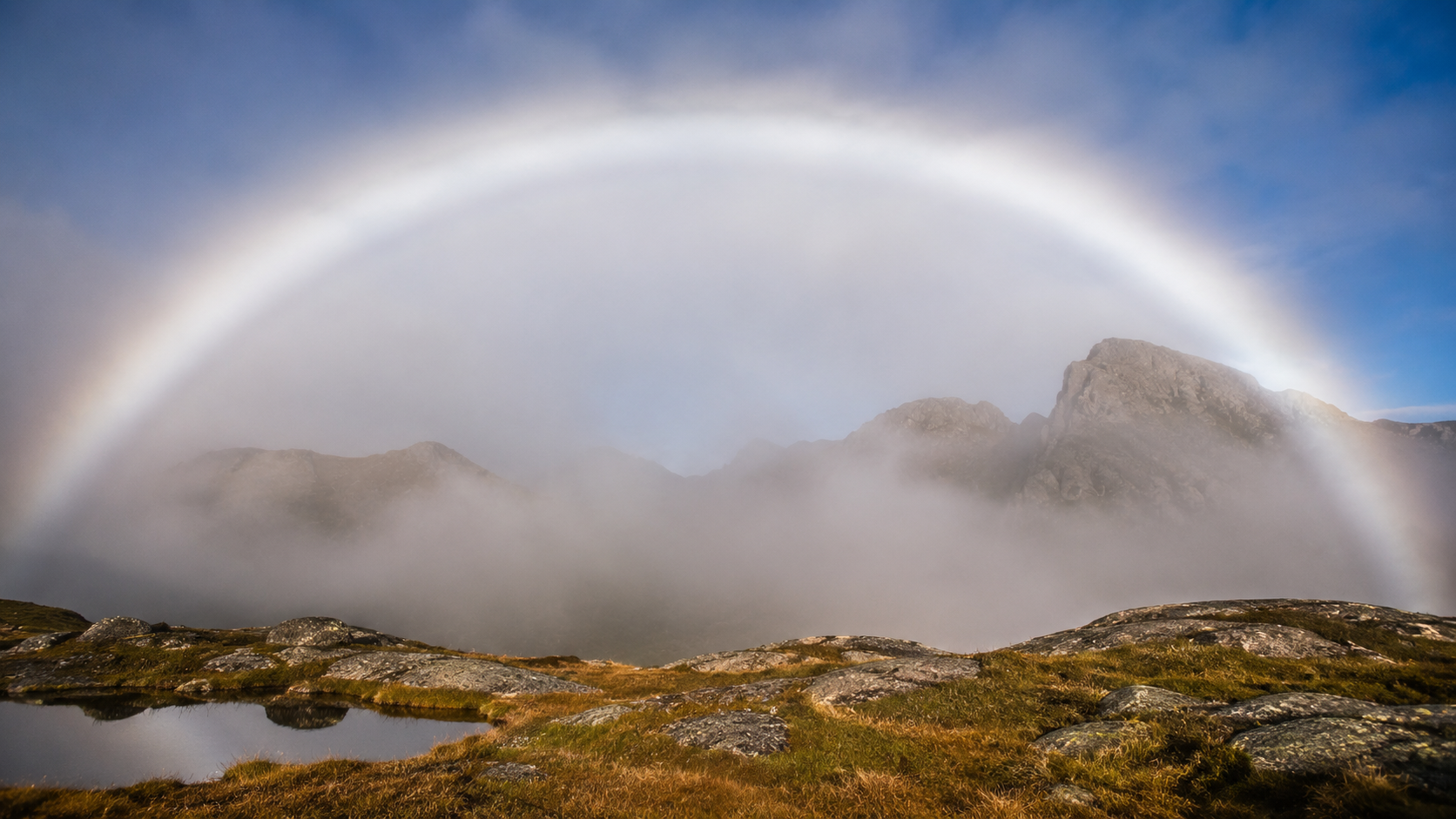 A realistic nearly white fogbow forming a huge pale arc above a mist-covered mountain valley, with only extremely subtle traces of color along its edges as sunlight breaks through the fog.